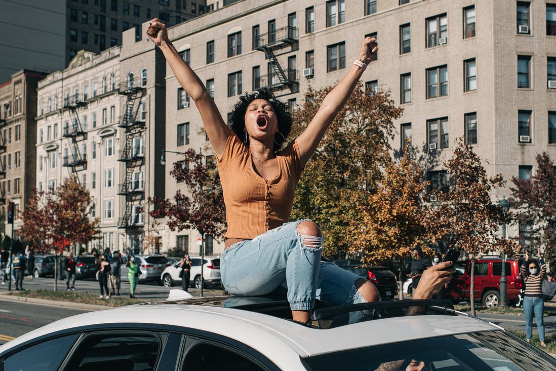A Black woman raises her arms in victory will sitting through a sunroof of a car